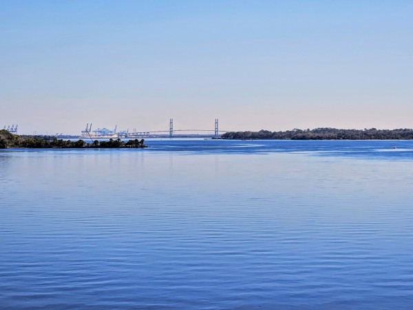 Very blue image. Beneath a light, clear blue sky, the calm blue waters of two rivers converge with an enormous suspension bridge and sea port cranes in the background.