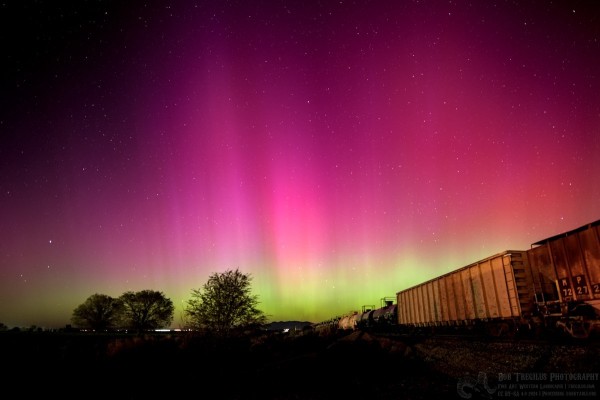 A color photo of the aurora. There are train tracks on the right side of the frame with yellow train boxcars on the tracks. There are three trees in the foreground left side and some lights in the far distance. The sky is green on the horizon, then a little bit of yellow, and finally a wide band of red. Stars are also visible in the sky.