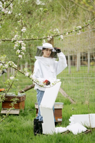 A coliur photograph of a young woman standing in a grassy field with cherry blossoms sweeping in branches in front of her. She is wearing black wellingtons, blue jeans and a white beekeepers jacket and hat. On hand is adjusting the brim of her hat, while the other holds some white material. 