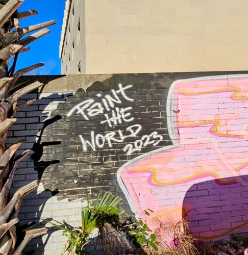 A parking lot facing wall for a small building is painted with the message "Paint The World - 2023" in white letters against a black background and nearby pink and yellow flags painted on the brick wall.