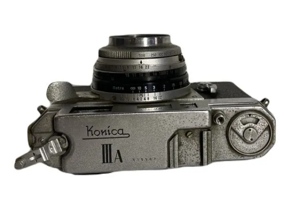 Photo of a vintage film camera from the top, the camera model name is more to the left side of the picture and it's "Konica III A"