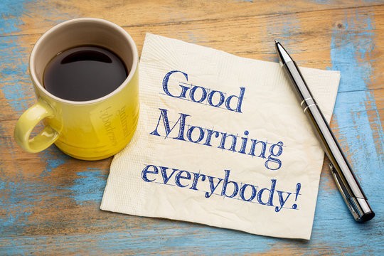 brown background with a yellow mug with black coffee in it, a white napkin with the words "Good Morning everybody" and a silver pen.