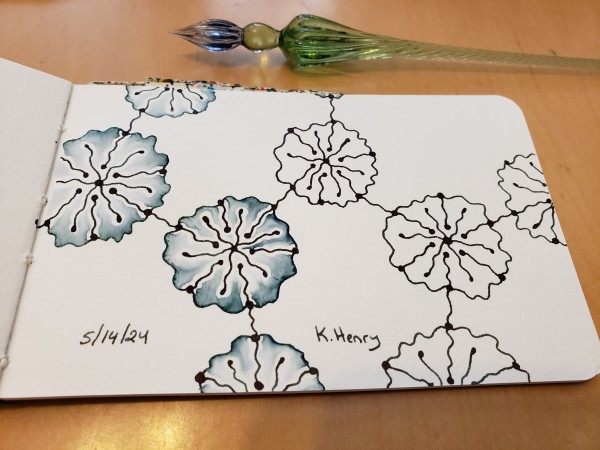 Hand drawn genertive/iterative art in ink on an open page of my sketchbook. The abstract pattern looks a bit like flowers on a network diagram. My glass dipping pen is next to my sketchbook.