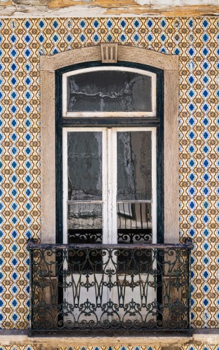 Color photo of a french door with window above it that opens onto a tiny balcony with black iron railing. The wall surrounding it is covered with decorative and colorful tiles. 