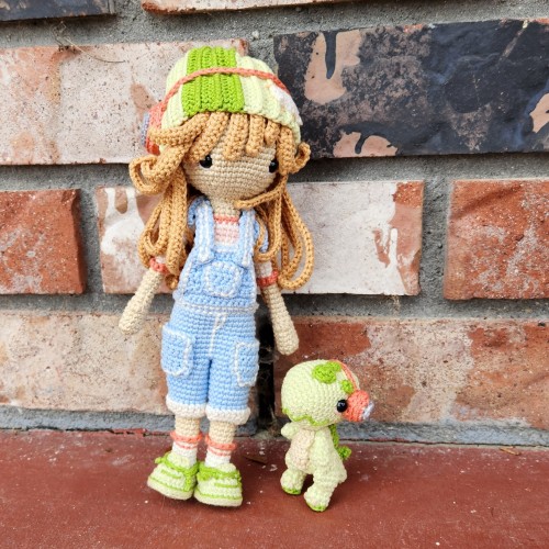 A crochet doll and a small dinosaur. Both are wearing headphones. The doll has blue jean overalls and a beanie to match their shoes and the dino.