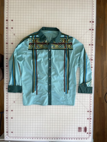 Front view of an azure button up, camp collared shirt. It has hunter green placket, collar, and cuffs. Brown, olive, teal, and sea glass colored mod work ribbon runs in parallel lines horizontally across the chest and shoulders. Brown, teal, olive, and turquoise ribbon fringe runs vertically from center shoulder to about six inches up from hem and is woven through the horizontal ribbon where they cross. The shirt’s buttons are blue and green abalone shell. The cuffs are vented with a green floral facing on the inner lap and snap French turnback. Snap is antique bronze. 