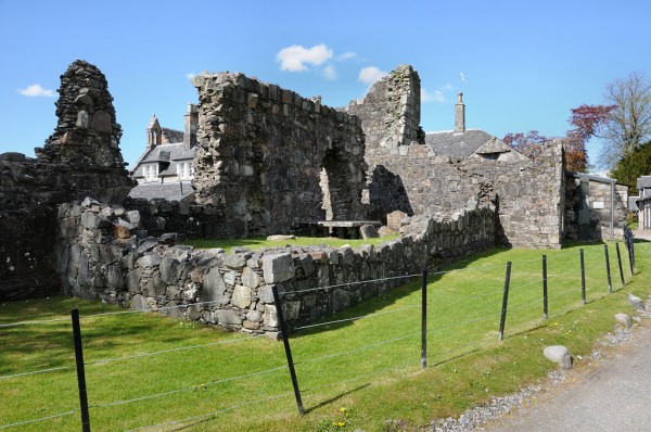 Ardchattan Priory Garden. The image shows a broad expanse of lawn sloping down from left to right with a large stone house at its upper part in the upper left of the frame. There’s a well-developed flower bed in the lower right of the frame and trees mid right. The scene is in sunshine.