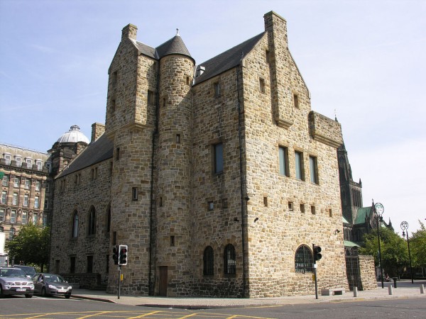 St Mungo Museum of Religious Life and Art. The image shows a corner view of a large castle-like building made of stone. In the foreground is a busy junction controlled by traffic lights. To its right is a glimpse of Glasgow Cathedral. The building is lit by sunlight coming from the right, leaving a long face on the left in shadow.