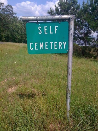 A very basic sign that says "self cemetery" in a field of grass with trees behind it