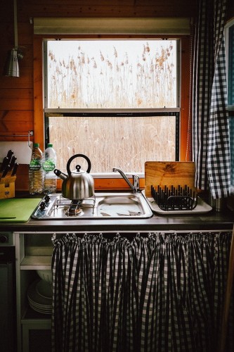 Eine gemütliche einfach Hausboot-Küche mit einer Spüle, einem Wasserkocher auf dem Gasherd, einem Schneidebrett und einem Messerblock an einem Fenster mit Blick auf hohes Schilf. Es gibt karierte Vorhänge und Wandpaneele aus Holz.