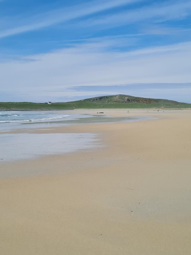 A sunny, sandy beach with green hills and a white farmhouse beyond