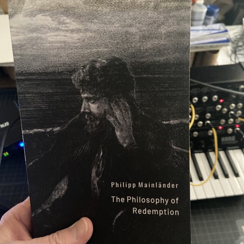 Photo of me holding a book. The book is titled “The Philosophy of Redemption” and the author is Philipp Mainländer. (The cover art of the book is black and white art, of a man looking to viewer left. His hand held up to the side of his head. There are clouds in the background.)

Behind the book is a synthesizer. 

