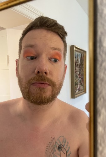 Redfern standing in front of a mirror, wearing eyeshadow that’s bright orange at the bottom and blending up to a soft pink. 