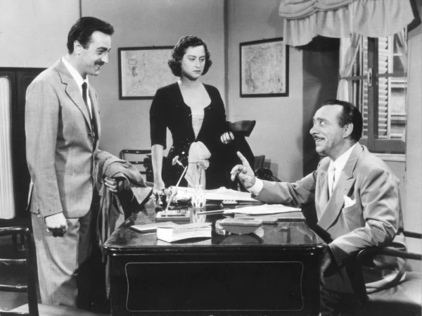  The image is a staged photograph featuring three individuals, likely from a film or television production. There's a man in the center who appears to be speaking, gesturing with his hands as he addresses another person seated across the desk from him. To his left, there is a woman wearing a black dress and a white blouse, leaning slightly towards the man at the table. On the right side of the frame, there's another man dressed in a light-colored suit and tie, looking at the first man with interest or engagement. All three characters are depicted in an indoor setting that resembles an office or study room, as indicated by the presence of a desk and various items on it such as books and papers. The image has a vintage appearance, suggesting it may be from an older film or TV show.