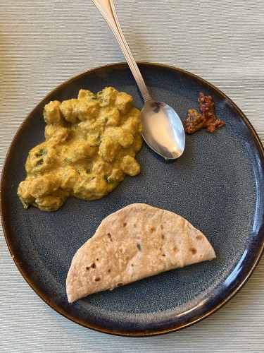 A blue plate with yellow gatte ki subzi, a roti, and a spoon with a small portion of a achar on a textured tablecloth.