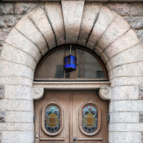 Brown door in a stone frame with two stained glass windows that look like eyes. There is a blue plaque that reads 15B in an Art Nouveau font set in the transom window.