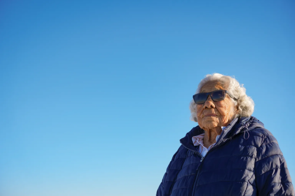 An elderly Native woman with short, wavy silver hair, a puffy jacket and sunglasses, against an expansive clear blue sky.