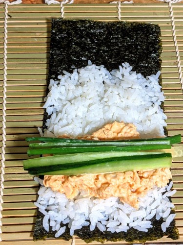 Bamboo rolling mat with a ½ sheet of nori with rice, spicy mayo salmon, cucumber and scallions on top ready to roll