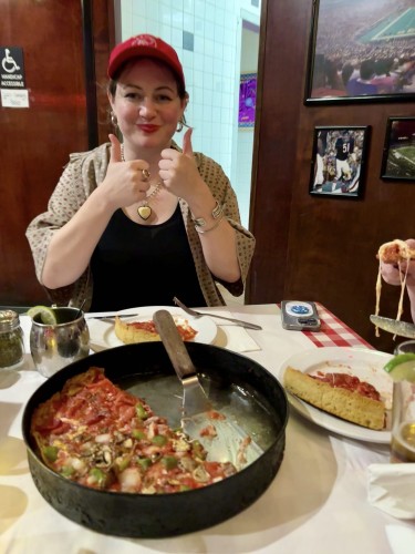 Pauline giving thumbs up at a table with a half-eaten deep-dish pizza, a slice of pizza on a plate, and framed sports photographs on the wall behind her.