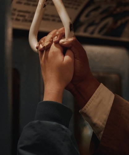 Photography. A color photo of two hands in a subway. A female white hand in a plain green jacket and a male black hand in a beige suit jacket and white shirt, holding each other and a subway handle loop. The photo symbolizes in a simple way the similarities between people beyond status, gender or skin color.