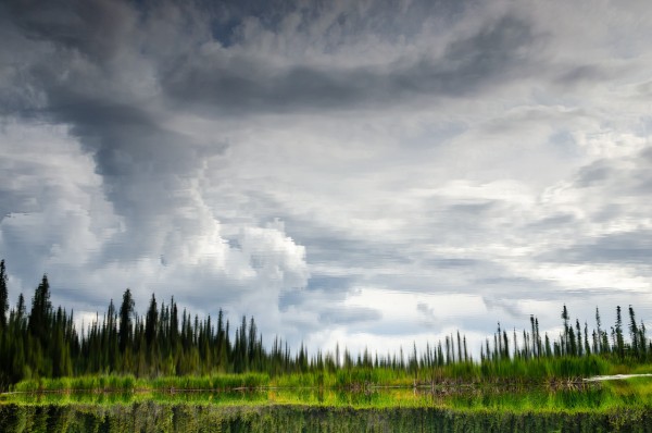 Inverted image of Ballaine Lake showing a vivid reflection of dark clouds and a dense forest. The sky, abundant with large, fluffy clouds, mirrors onto the water's surface, creating a striking and topsy-turvy visual effect. The upper portion of the image features the reflection, while the lower portion displays the actual trees along the lake's edge, subtly blurred. This scene captures a late summer day at Ballaine Lake in Fairbanks, Alaska, emphasizing the surreal beauty of reflected landscapes.