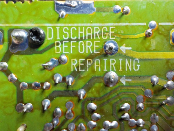 The back of the PCB of a power supply, with two large pads, and arrows pointing at them, and a text written in capital letters saying: Discharge before repairing.