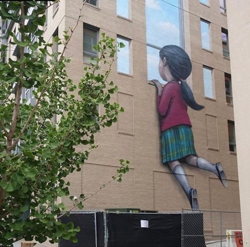 a photo of a building, in between the windows is a large painting of a young girl holding onto the bottom of a windowsill, her feet dangling, there are blue clouds and sky through the large window she is looking through 