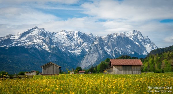 A picturesque scene of a rural landscape featuring a group of buildings surrounded by a vast field. In the background, majestic mountains rise up against the sky, creating a stunning backdrop.  Two prominent objects stand out in the image: a tree in the foreground and a house slightly off-center. The composition evokes a sense of tranquility and natural beauty, with hints of a springtime atmosphere. This serene setting captures the essence of a peaceful countryside retreat, with a touch of alpine charm.