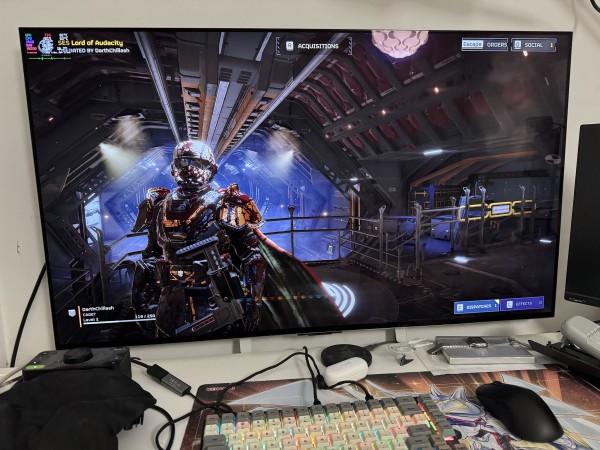 Computer monitor displaying a futuristic video game character in armor standing in a sci-fi environment, with gaming peripherals on a desk in the foreground.