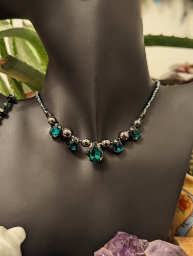 handmade dark green gothic necklace with loads of dark green drop pendants in varying sizes and hematite styled beads and glass dark blue beads
on black doll with plants and crystals around
