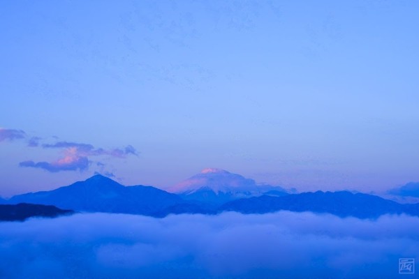 a pink alpenglow Mt Fuji with some clouds around it, but a soid blanket of clouds beneath it at the bottom of the photo