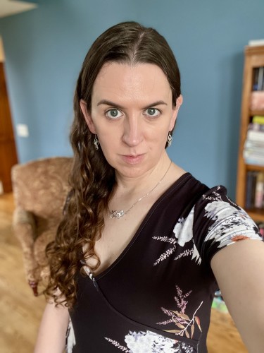 Selfie of a woman with long curly brown hair. She wear a low cut black floral top, a necklace, and earrings. Her hair falls forward over one shoulder.