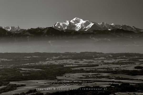 Black and white photograph showing a panoramic view of the Mont Blanc massif. In the foreground are fields, woods and buildings, and just below the massif is a light mist over Lake Leman, which passes smoothly and ends just below the summit of Mont Blanc.