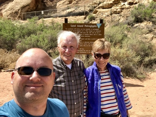 Three people posing for a selfie with a sign that reads "Sand Island Petroglyphs" in the background. A fenced-off rocky cliff area is visible behind them.
