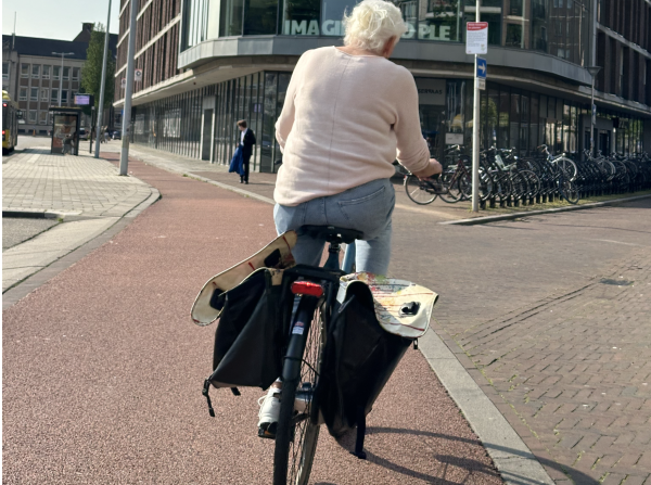 An old person riding a bike along a smooth bike path. She is turning right into the street that has many bikes parked along it. In the background a man walks toward a bus stop. There are no cars visible.