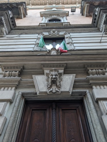 An ornate carved stone doorway with a hardwood dark brown door. Over the door is an open-mouthed, howling gargoyle. Overthat is a pair of Italian tricolor flags framing a window. More windows and stonework are visible above it.