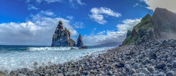 Das Bild zeigt die in der Mündung der Ribeira da Janela im Meer stehenden typischen Felsen vor blauem Himmel mit weißen Wolken. Am rechten Bildrand ragt die Insel hoch auf und im Hintergrund lässt sich das andere Ende der Insel am Horizont erahnen.
