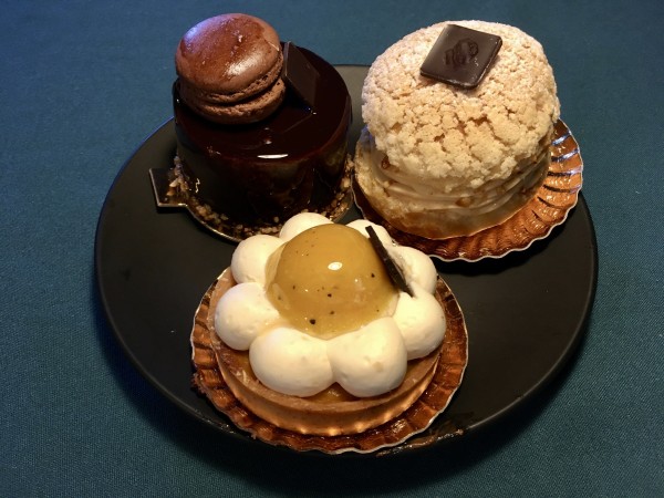 Sur une assiette à dessert noire, 3 gâteaux individuels : une tartelette décorée d'une fleur aux pétales en crème à la noix de coco et au cœur passion, un entremets couvert d'un glaçage chocolat et décoré d'un macaron, et un chou façon Paris-Brest.