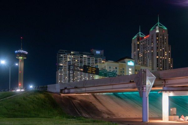 Paysage de nuit, photo prise en contre-bas d'une autoroute à proximité d'un pont, d'où on voit dépasser un panneau où il est inscrit : "Arrivez tôt, restez sur place, repartez tard". A l'arrière plan des gratte-ciels illuminés et deux hôtels. 