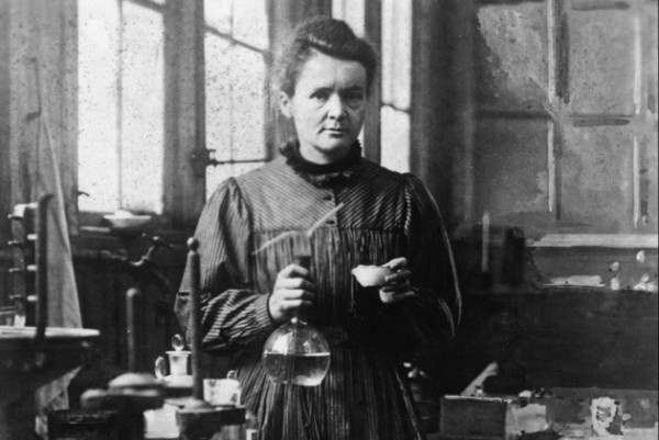Marie Curie in a posed photo in her lab. She's a white woman with dark hair, holding a glass flask and a metal or ceramic dish.