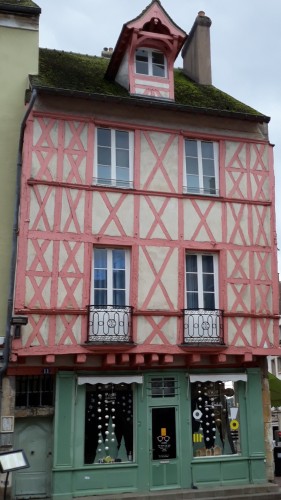 Une maison de ville à deux étages plus une soupente, avec des colombages rose bonbon. Le rez-de-chaussée est occupé par un magasin dont l'entourage en bois de la devanture  est peint en vert.