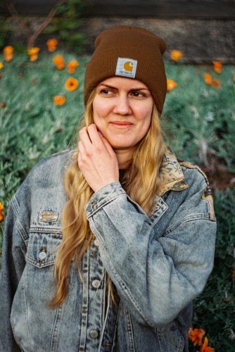 A blonde-haired woman wearing a brown winter cap and gray denim jacket, looks to the right, holds her left hand to her neck. The background shows orange flowers and gray-green leaves, the background swirls.