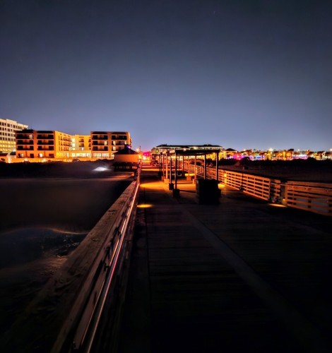 Beneath a clear night sky an orange glow of night lighting illuminates a big wooden pier extending out into the Atlantic's waters, with the slow, calm surf seen washing up on the dark beach in front of a coast lined with hotels and other properties also glowing with colorful night lighting.