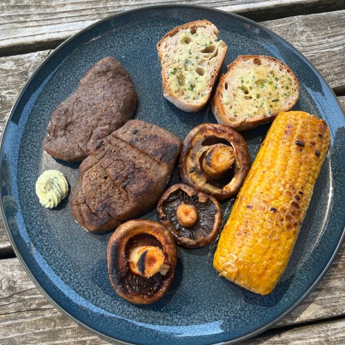 Ein dunkler Teller mit gegrillten Seitansteaks, Champignons, Maiskolben und Baguette mit veganer Kräuterbutter.