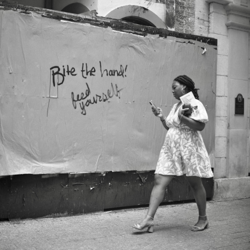 A black and white film photo. A woman walks passed graffiti that states “Bite the hand. feed yourself” written a temporary wall in downtown Austin Texas. Tuesday, April 30, 2024.