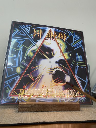 A Def Leppard vinyl record album cover displayed on a wooden stand, featuring with geometric shapes and neon-like accents. Text on the album includes "Def Leppard" and “Hysteria”