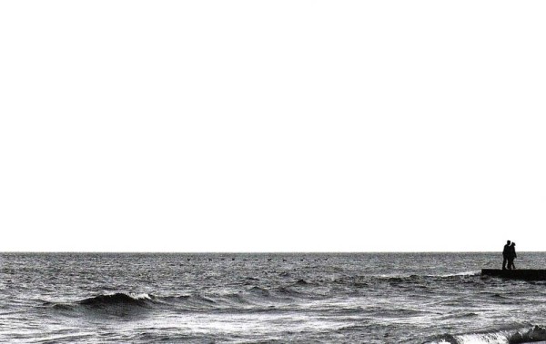 Imagen en blanco y negro dividida en dos horizontalmente por la línea del horizonte en un lago. El cielo ocupa unas tres cuartas partes de la imagen. En el extremo derecho, el borde de un rompeolas sobre el que hay dos personas (siluetas en negro) caminando juntas.