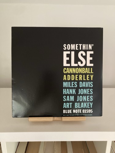 Vinyl album cover for "Somethin' Else" by Cannonball Adderley featuring Miles Davis, Hank Jones, Sam Jones, and Art Blakey displayed on a shelf. The cover is plain black with white, green and blue letters.