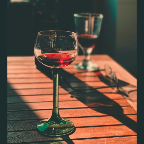 A glass of red wine is standing on a brown table. In the background you see glasses on the right hand side and another glass of red wine.