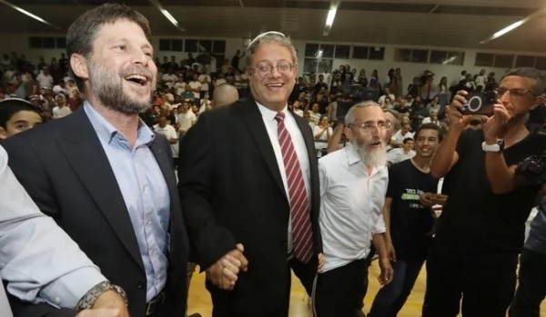 Israeli ministers Bezalel Smotrich and Itamar Ben-Gvir, in 2022 circle dancing in a crowded room full of religious nationalists Israelis, with onlookers and a person taking a photo in the background.